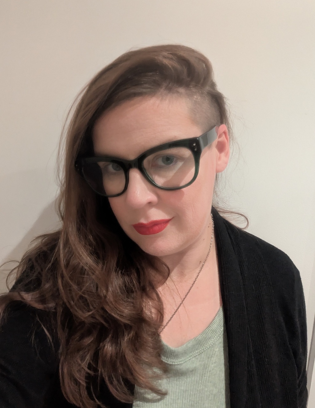 JC Darcy, a white femme presenting non binary person with long brown hair in an undercut and black glasses smiles at the camera with a cocked head.