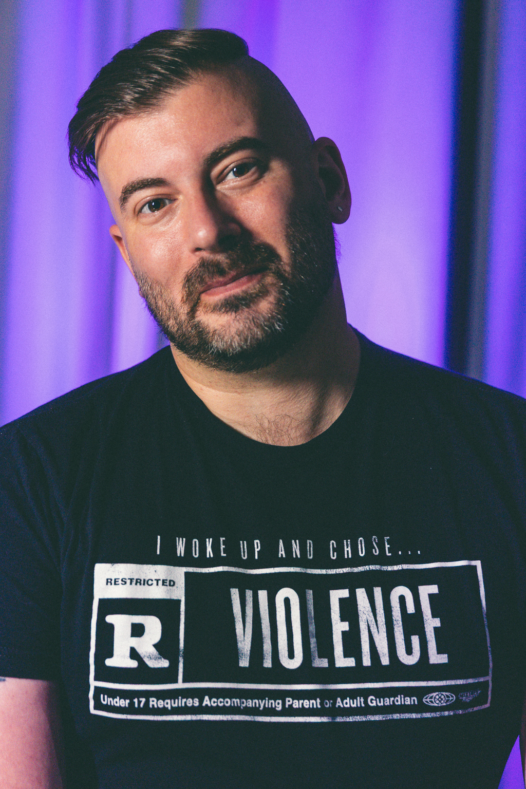 Eric Crumrine, a tanned white masc presenting person with an undercut and a beard, smiles at the camera. He is wearing a shirt that reads "I woke up and chose... followed by a film classication box stating rated R for Violence.