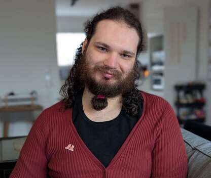 Kieran, a white masc presenting non-binary person with curly brown hair and a beard, looks at the camera and smiles softly. They are wearing a black top and red cardigan.